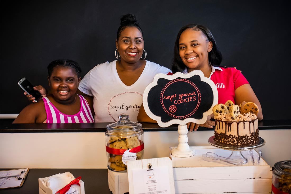 three women standing behind a counter with a cake and cookies.